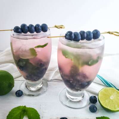 blueberry mojito in wine glass with fresh blueberries and mint, with sliced limes on wood backdrop
