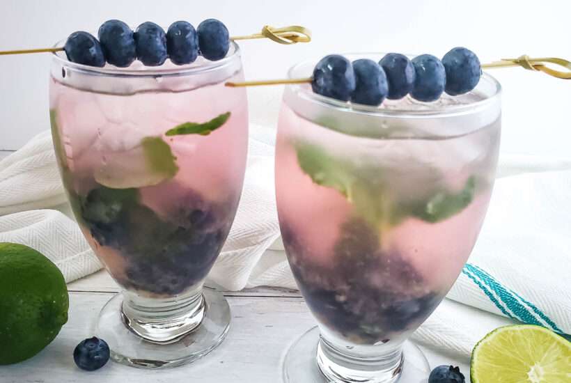 blueberry mojito in wine glass with fresh blueberries and mint, with sliced limes on wood backdrop