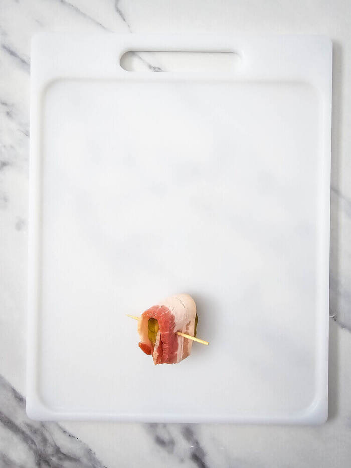 pickle wrapped in bacon on a cutting board with a tooth pick
