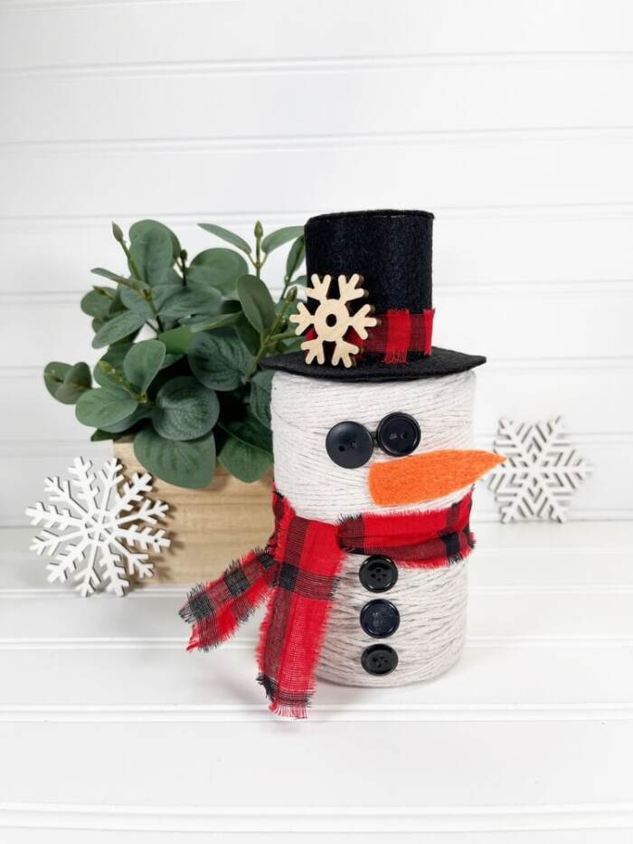 Finished DIY Snowman String Ball Craft made from white string with a black felt top hat, red plaid scarf, button details, and orange felt carrot nose displayed on a white shelf.