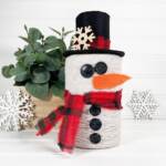 Finished DIY Snowman String Ball Craft made from white string with a black felt top hat, red plaid scarf, button details, and orange felt carrot nose displayed on a white shelf.