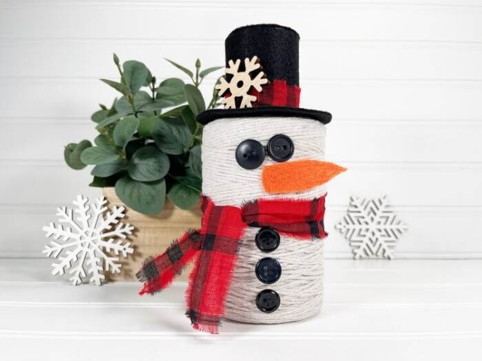Finished DIY Snowman String Ball Craft made from white string with a black felt top hat, red plaid scarf, button details, and orange felt carrot nose displayed on a white shelf.