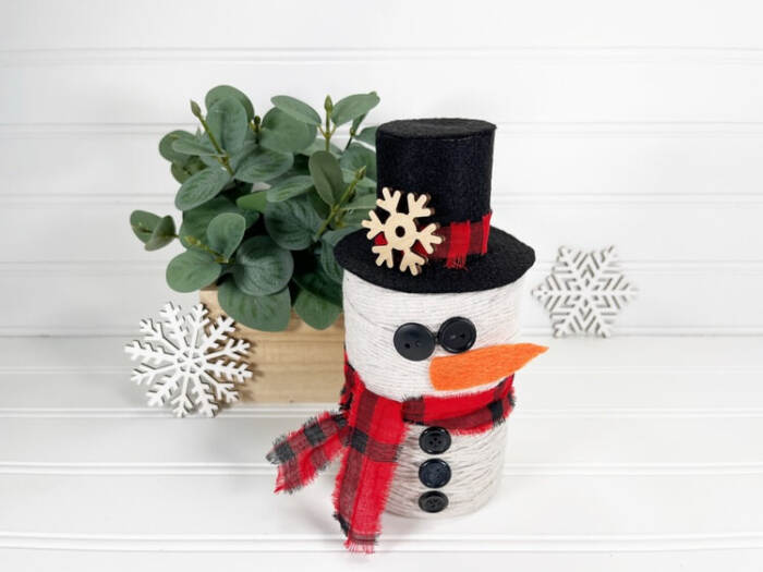Finished DIY Snowman String Ball Craft made from white string with a black felt top hat, red plaid scarf, button details, and orange felt carrot nose displayed on a white shelf.