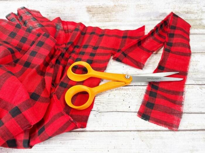 Red and black plaid fabric being cut into strips with scissors to make a snowman scarf.