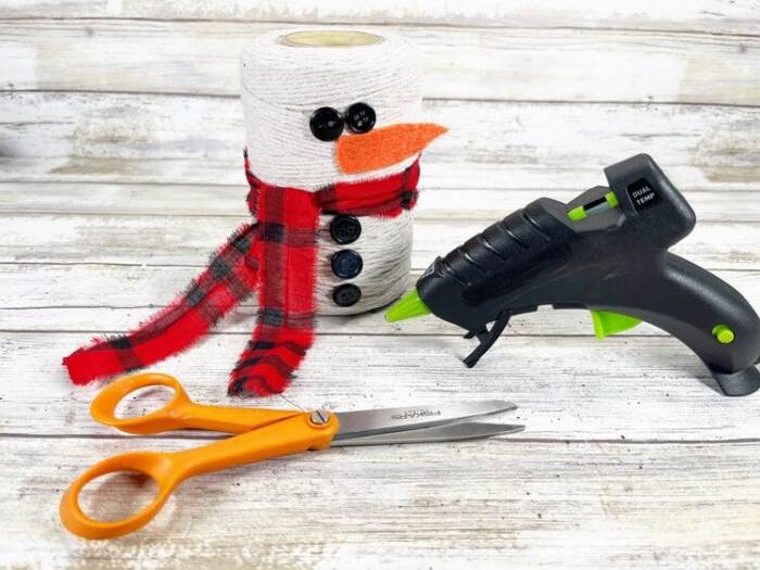 Snowman with plaid scarf and black buttons, shown with scissors and a glue gun.