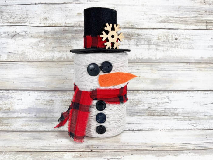 Finished snowman decoration standing upright with hat, scarf, buttons, and carrot nose on a wooden background.