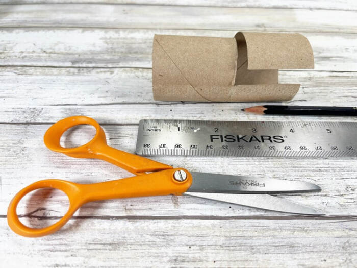 Cardboard toilet paper tube cut to size with scissors, ruler, and pencil on a wooden surface.