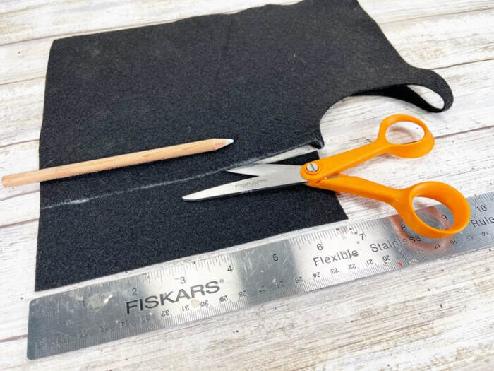 Black craft felt being measured and cut with scissors, ruler, and pencil on a wooden work surface.