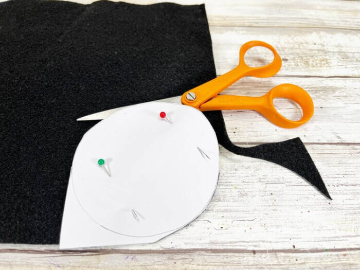 Paper snowman hat pattern pinned to black craft felt with scissors beside it on a wooden surface.