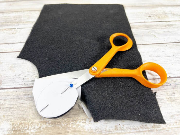 Small paper hat pattern pinned to black craft felt being cut out with scissors on a wooden surface.