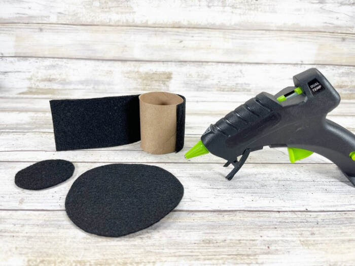 Black felt hat pieces and a cardboard toilet paper tube arranged with a hot glue gun on a wooden surface, ready for assembly.