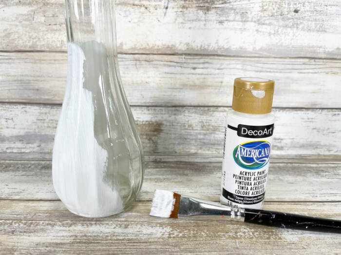 Glass vase partially painted white with white acrylic paint, paintbrush, and paint bottle on a wooden surface.