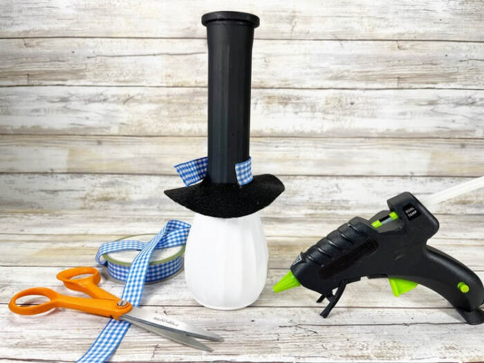 Snowman vase painted white and black with blue gingham ribbon, scissors, and glue gun on a wooden surface.