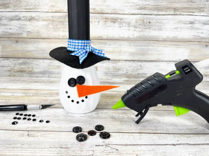 Snowman Vase Craft with black button eyes, rhinestone mouth, and orange felt carrot nose being attached.