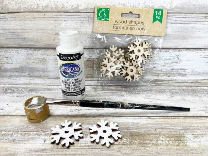 White acrylic paint, paintbrush, and wooden snowflake shapes used for Snowman Vase Craft decoration.