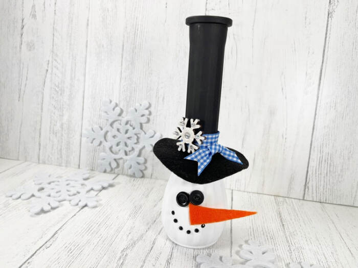 Snowman Vase Craft made from a painted glass vase with a black top hat, orange felt carrot nose, button face, and winter decor background.