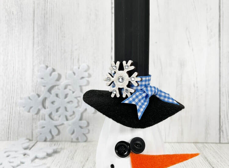 Snowman Vase Craft made from a painted glass vase with a black top hat, orange felt carrot nose, button face, and winter decor background.