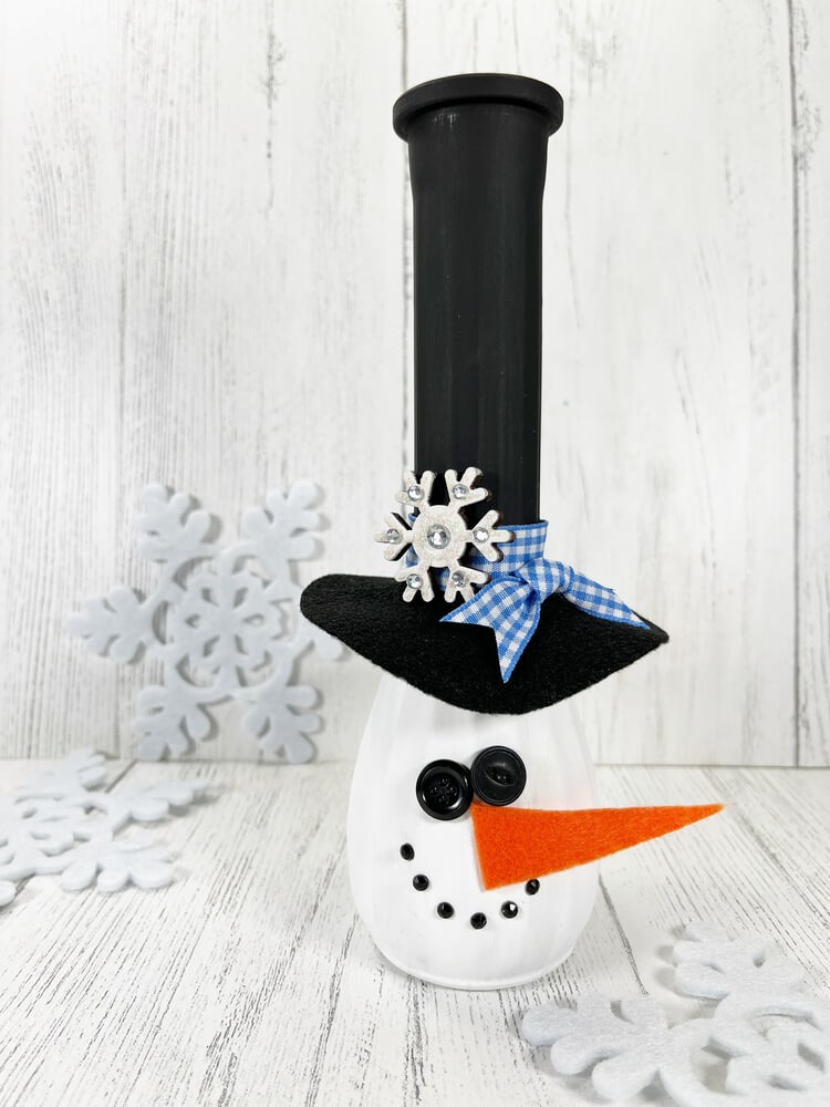 Snowman Vase Craft made from a painted glass vase with a black top hat, orange felt carrot nose, button face, and winter decor background.