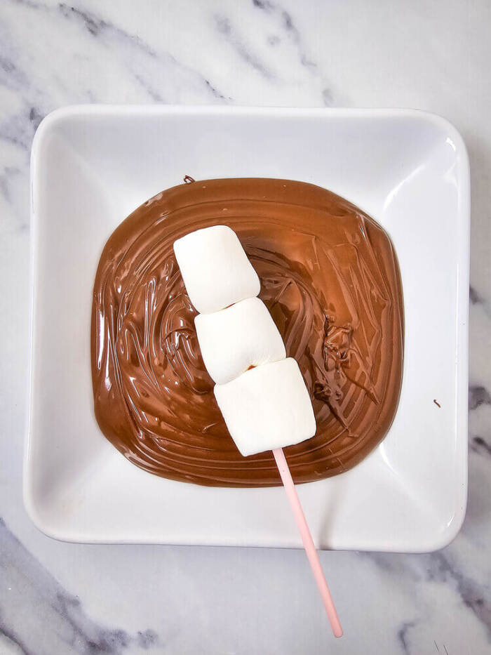 Three marshmallows on a pink stick being dipped into melted chocolate in a white square dish.