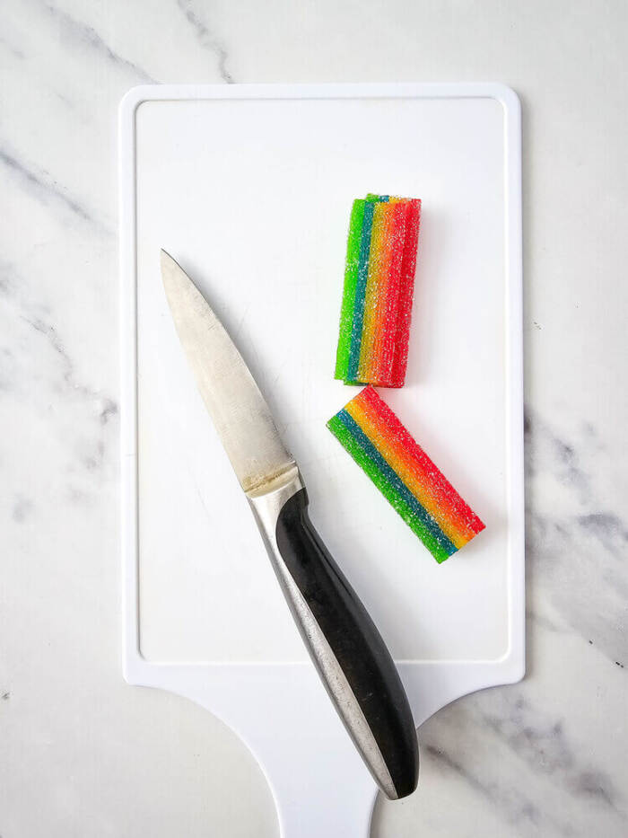 small white cutting board with a kitchen knife and two stacked pieces of rainbow sour candy strips on it.