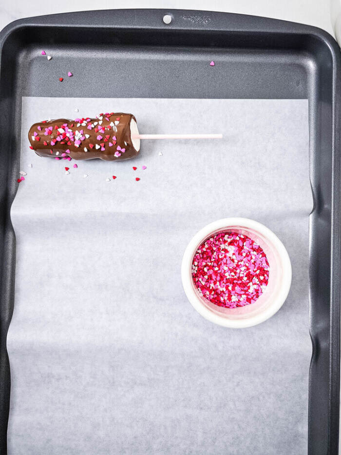 Chocolate-covered marshmallow pop on a baking tray with a bowl of pink and red heart sprinkles