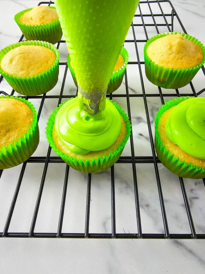 Piping bright green frosting from a piping bag onto plain cupcakes that are on a cooling rack.