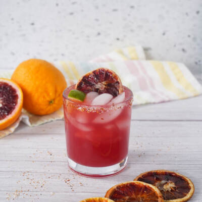 Blood orange margarita in a short glass with ice, lime wedge, and dried blood orange garnish on a white table.