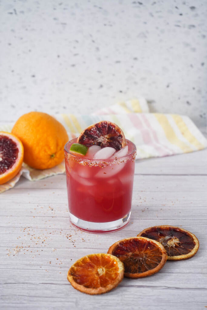 Blood orange margarita in a short glass with ice, lime wedge, and dried blood orange garnish on a white table.
