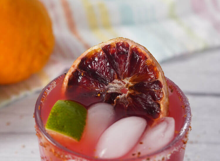 Close up of blood orange margarita showing ice, lime wedge, and dried blood orange slice garnish.