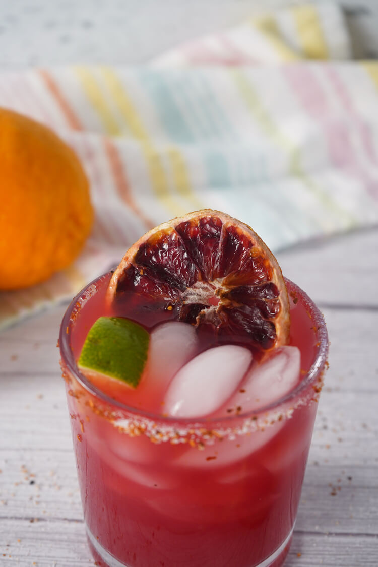 Close up of blood orange margarita showing ice, lime wedge, and dried blood orange slice garnish.
