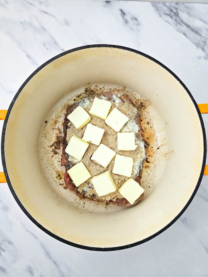 Raw dutch oven mississippi pot roast topped with seasoning and butter.