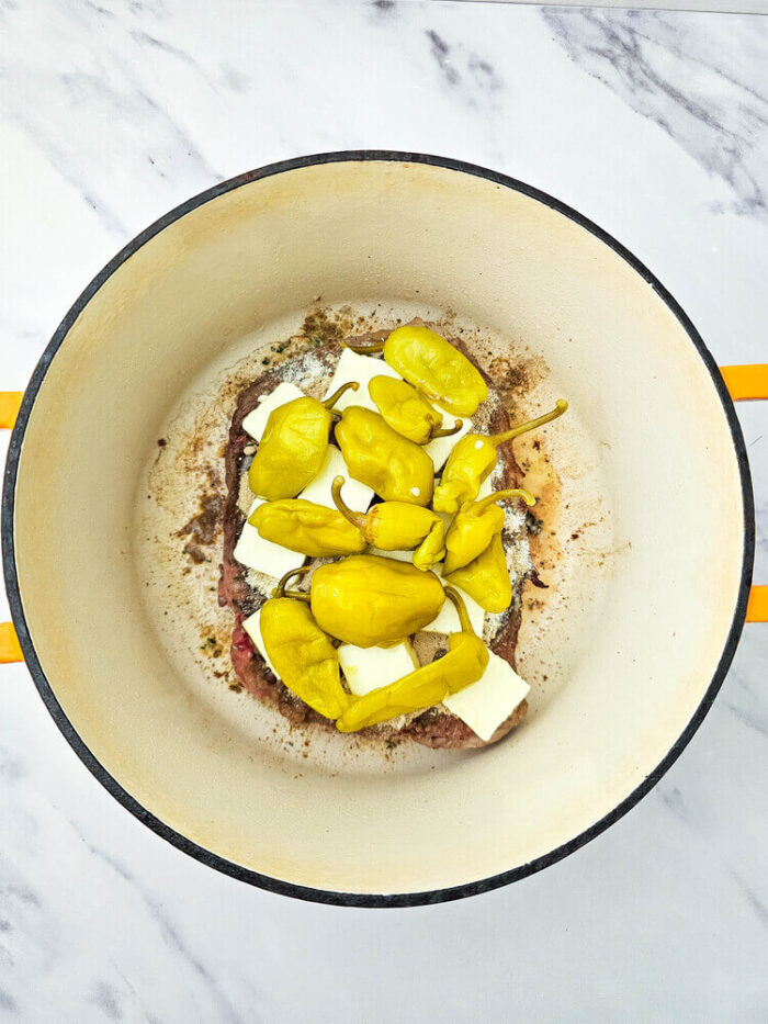 Raw dutch oven mississippi pot roast covered with seasoning and topped with butter and peppers.
